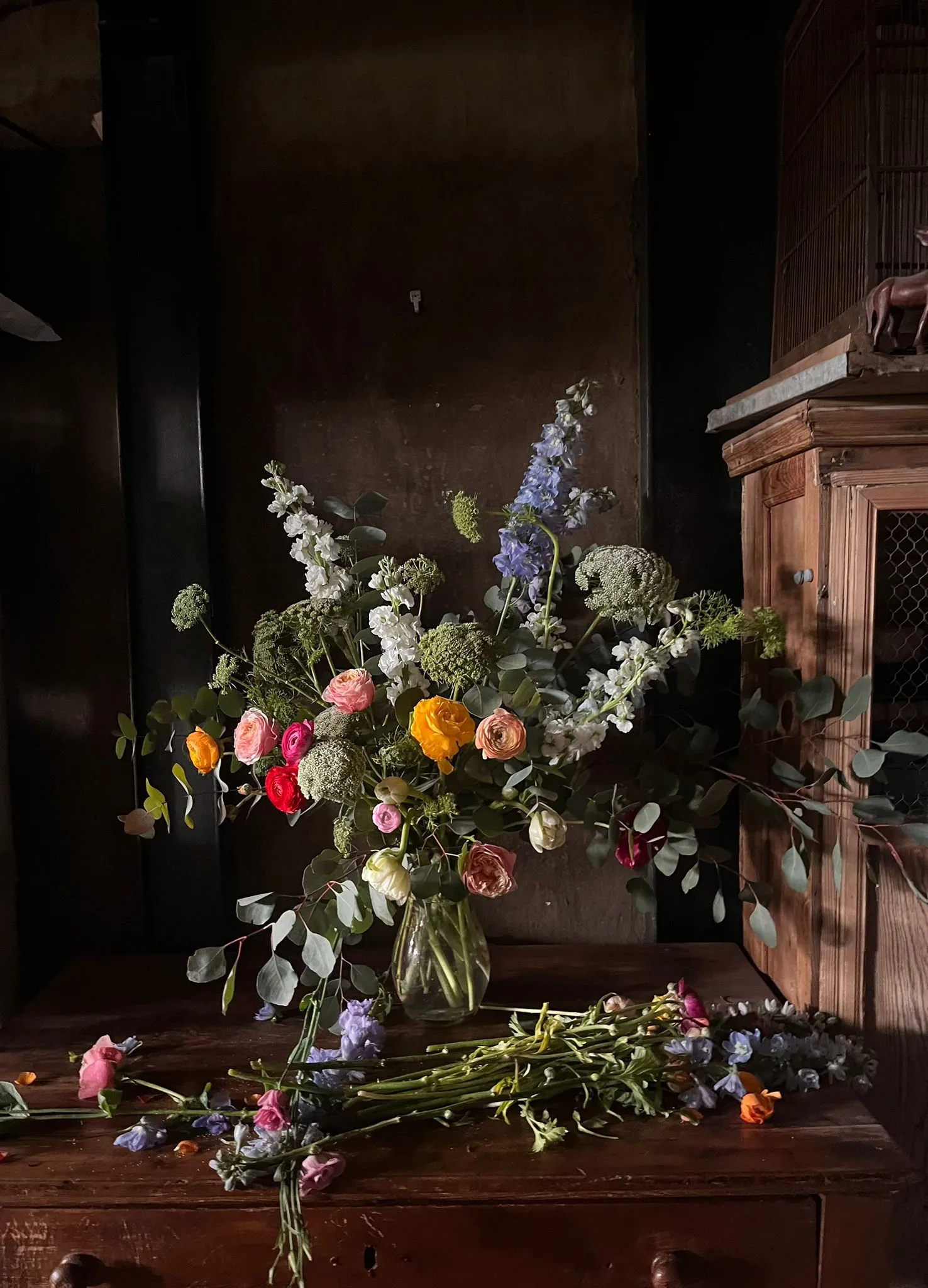 Ramo de ranúnculos, delfinium y eucalipto en jarrón de cristal sobre mesa de madera oscura